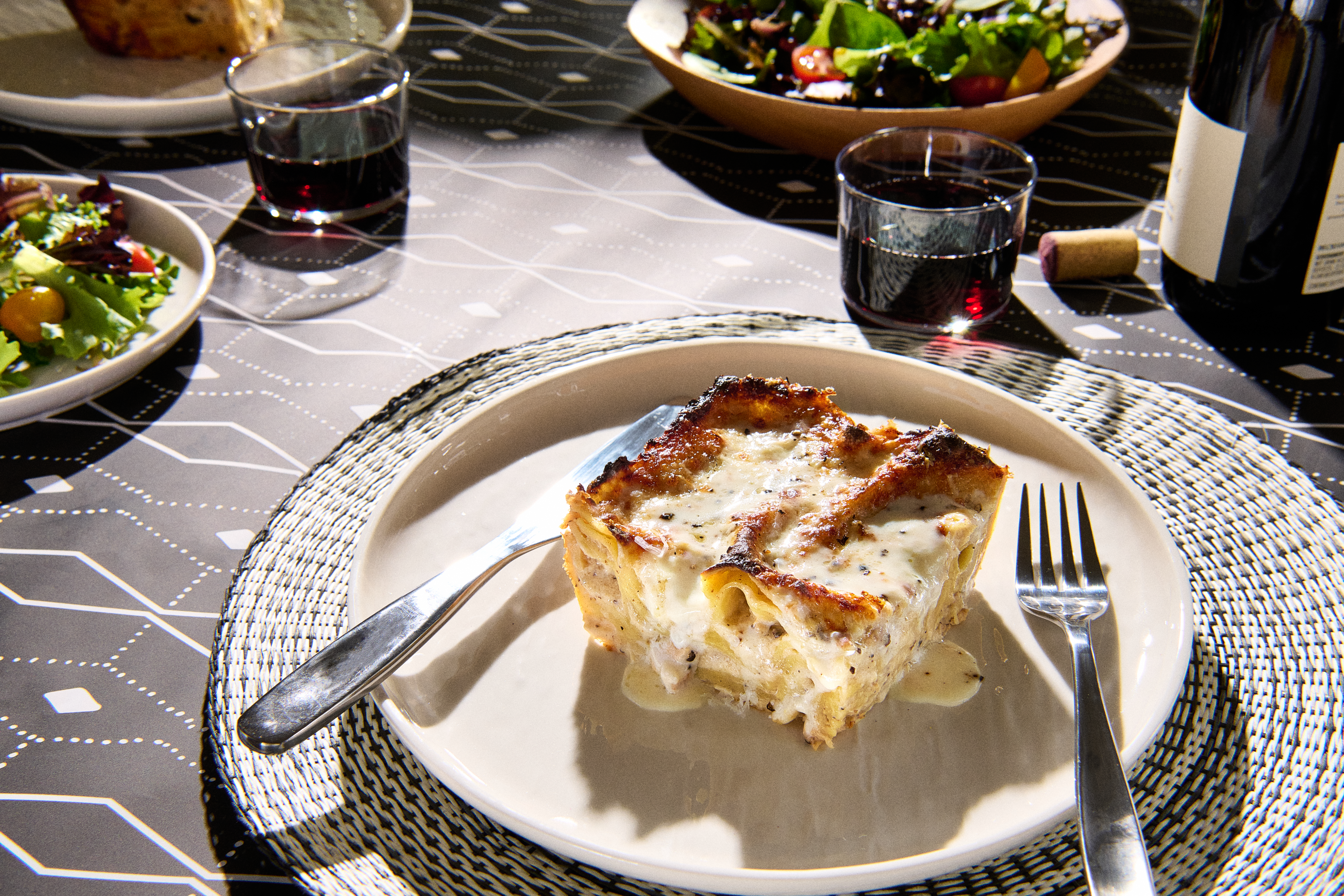 A square of lasagna served on a plate, with glasses of red wine and a salad nearby.