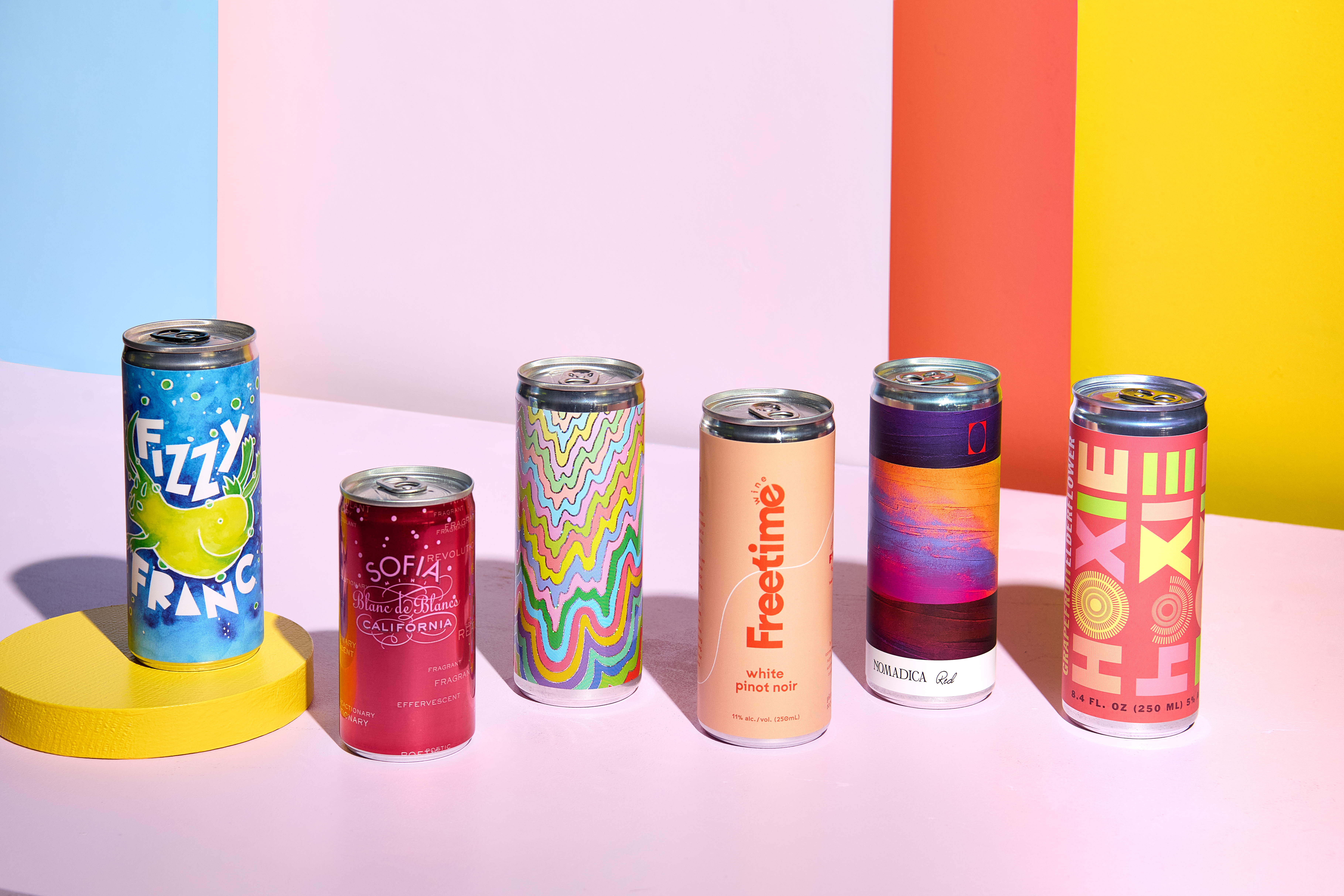 Six colorful wine cans sit on a pink table.