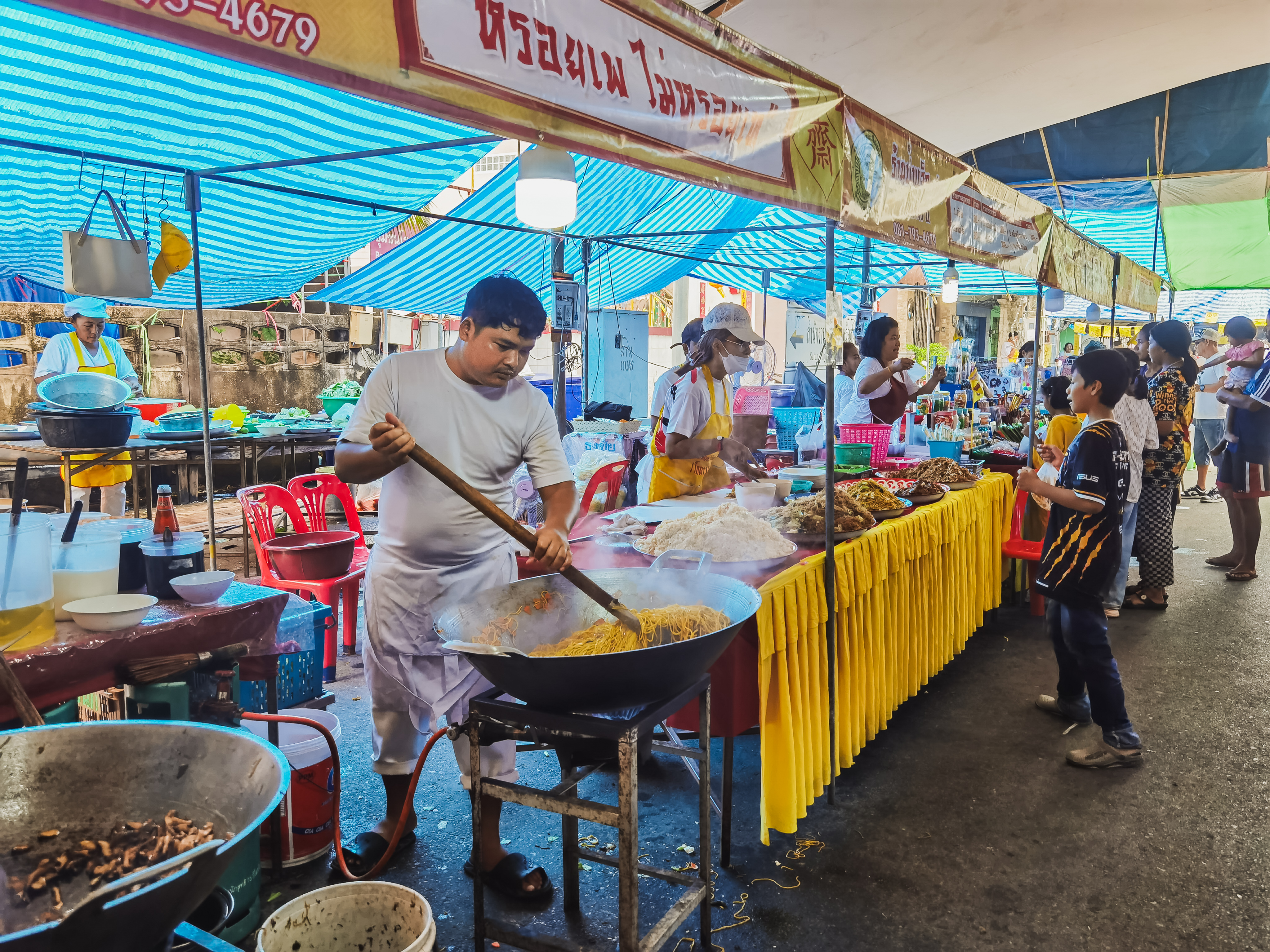 A food vendor stirs a large wok of noodles with a long poll.