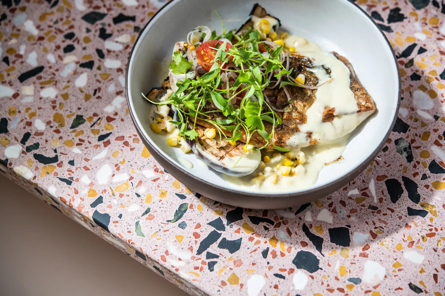 A white bowl of a large filet of skin-on Gulf fish, surrounded by corn maque choux and clams and topped with micro-greens.