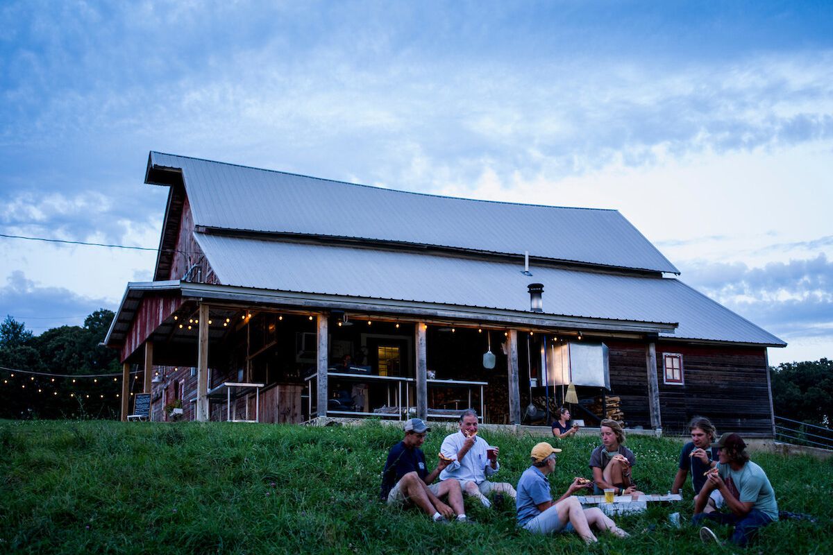 A small farm building underneath a partly cloudy sky at dusk, with people gathered on the grass and eating.
